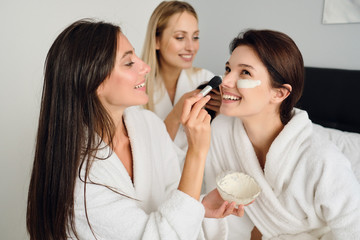Obraz na płótnie Canvas Three young beautiful women in white bathrobes sitting on bed happily making cosmetic mask together in modern hotel