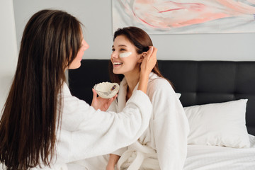 Obraz na płótnie Canvas Two young beautiful women with dark hair in white bathrobes sitting on bed happily making cosmetic mask together in modern hotel