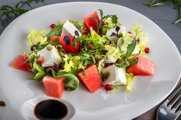 Summer salad with watermelon and feta cheese salad in a white plate. Brown tablecloth, spoon, fork on the grey background. Top view, closeup