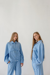 two pretty young twin sisters with long blond hair posing on white background in oversize clothes. Fashion photoshoot