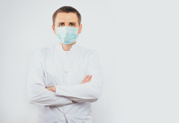 A man wearing a white coat and a mask on face has crossed arms on white background. Medical and pharmaceutical concept. A man dressed like a doctor. Preparation for the examination, health problems.