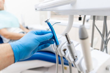 Close-up hand of dentist in the glove holds dental high speed turbine. The patient in blue chair at the background. Office where dentist conducts inspection and concludes. - obrazy, fototapety, plakaty