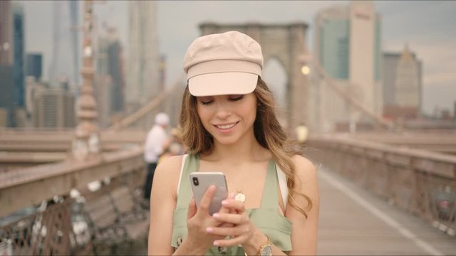 French lady walks by Brooklyn Bridge and chat with friends on a phone, sharing emotions and impressions, traveler's life, first time in New York, visiting destinations.