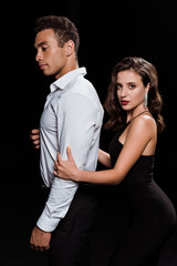 elegant woman hugging handsome mixed race man looking at camera isolated on black