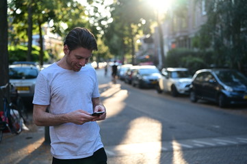 Spontaneous young man smiling and texting his friend on his mobile phone while standing in a ambient street