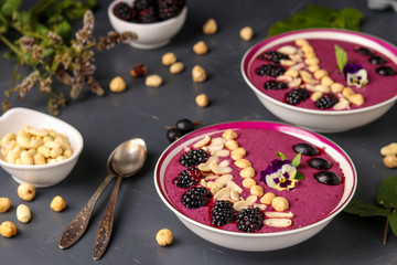 Healthy breakfast smoothie bowl with blackberries, gooseberries, peanuts, hazelnuts, decorated with flowers on a dark background