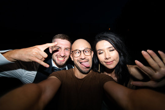 Smiling man with excited wedding couple taking selfie on black background at night