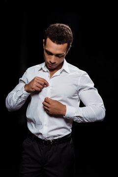 handsome bi-racial man touching and looking at white shirt isolated on black