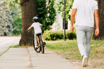 back view of kid riding bicycle while father walking after son