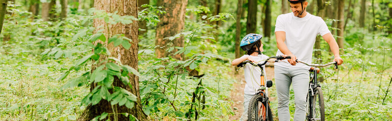 panoramic shot of father and son walking with bicycles in forest