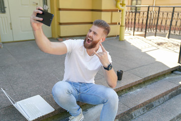 young good looking bearded guy is sitting outdoors on stairs in front of his house with laptop, making selfie and showing rock sign. Wearing white shirt and jeans