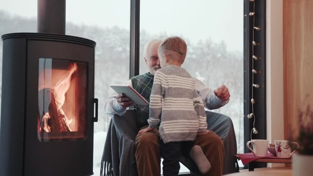 Happy grandfather and grandson using digital tablet for surfing internet and playing game near the fireplace at home grandpa adult grandchild child childhood communication computer slow motion