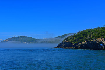 Fototapeta premium Morning fog hugs the coast of Deception Pass as it enters the Salish Sea in Washington State