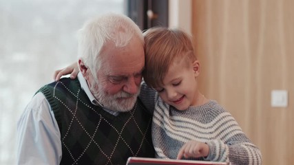 Smiling grandfather and grandson using digital tablet for surfing internet and playing game near the fireplace at home grandpa adult grandchild child childhood communication computer - Powered by Adobe