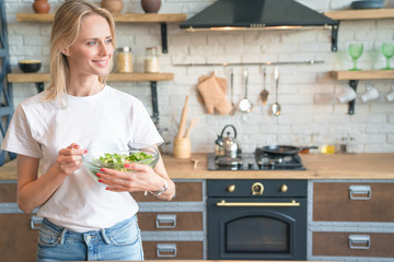 beautiful young smiling woman holding green salad in the kitchen. looking sideways. Healthy food. vegetable salad. Diet. Healthy lifestyle. cooking at home. Wearing white shirt and jeans.