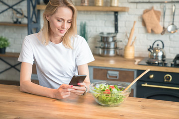 beautiful young woman using cell phone while making salad in the kitchen. Healthy food. vegetable salad. Diet. Healthy lifestyle. cooking at home. Wearing white shirt and jeans.
