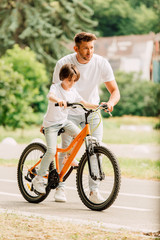 full length view of father looking forward and helping son to ride on bike while son sitting on bicycle