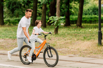 Obraz premium full length view of father pushing sit of bike and running after son while kid riding bicycle
