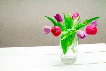 Copy space with beautiful pink and purple tulips with green leaves bouquet in a transparent glass vase on white wooden table on gray background