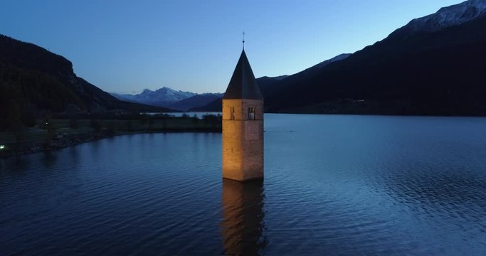 Aerial: Slow Pan of the Bell Tower of Lake Reschen at Night with Mountains in the Background - South Tyrol, Italy