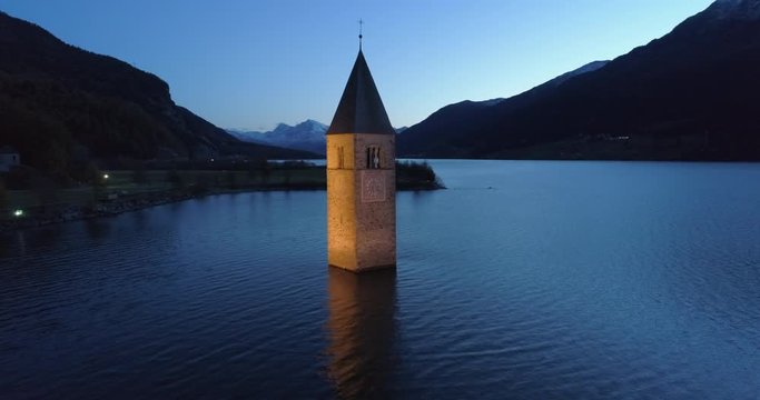 Aerial: Slow Pan of the Famous Lake Reschen Bell Tower at Night Between Hills and Mountains - South Tyrol, Italy
