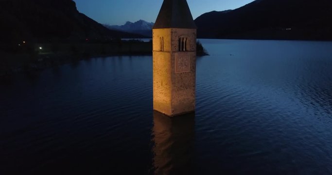 Descending Aerial: The Brightly Lit Bell Tower of Lake Reschen at Night Before Dark Hills and Mountains - South Tyrol, Italy