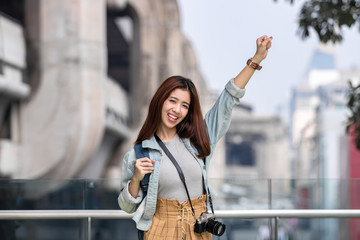 Cheerful young Asian woman tourist raising hands in urban city. Travel and vacation concept.
