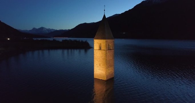 Descending Aerial: The Picturesque Bell Tower of Lake Reschen at Night Highlighted Against the Sky and Dark Hills - South Tyrol, Italy