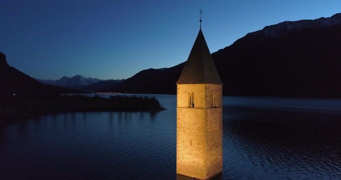 Aerial of the Illuminated Lake Reschen Bell Tower and Dark Hills and Mountains in the Far Distance - South Tyrol, Italy