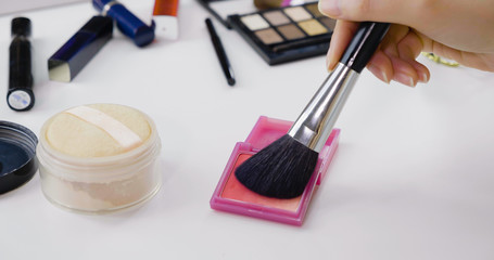 Close up of woman hands holding makeup brush applying pink blush on white vanity table. Female using cosmetics products for make up. messy dressing table with power and palette indoors.