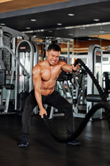 Fototapeta na wymiar Strong shirtless Vietnamese man doing exercises with battle rope during functional training in gym
