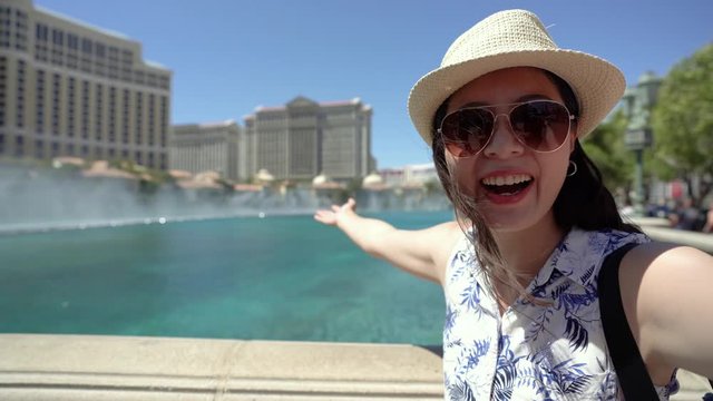 Young beautiful girl traveler standing in city near fountains outdoor summer. happy female backpacker waving hands while talking on video phone call. woman tourist showing camera view in background