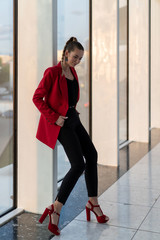 beautiful European girl in red jacket, heels and black trousers posing leans on panoramic glazing business center. Full height. Vertical photo.