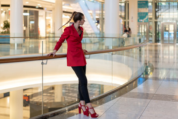 young white girl in red jacket, heels and black trousers leans on railing in mall and looks away. Full height.