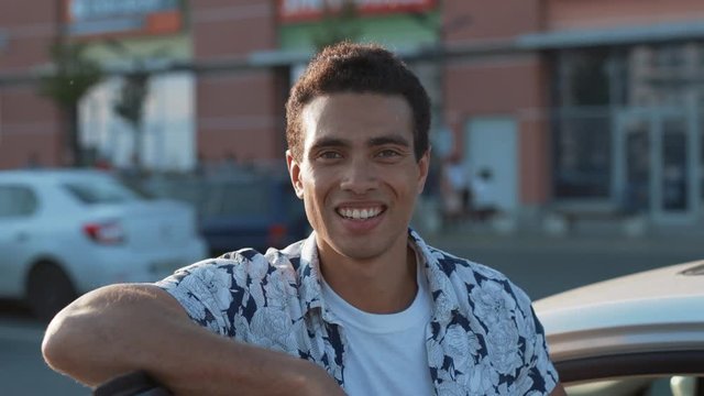 Outdoor portrait of nice-looking positive guy in stylish outfit leaning on modern car door. Close-up handsome young man posing outdoors against modern shopping mall.