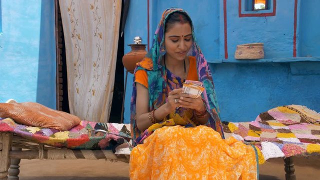 Happy Indian woman calculating her last month's savings - future investment. Village life. Young attractive villager in traditional saree happily sitting on a charpai  counting her savings of the p...