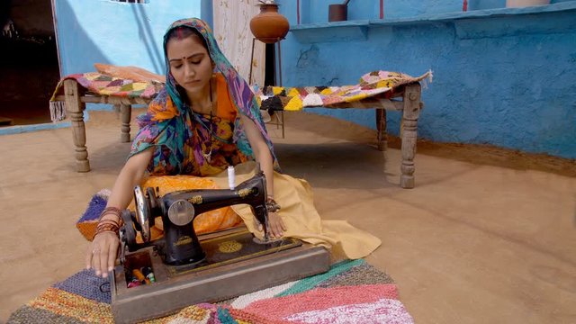 Indian village lady stitching her clothes on a sewing machine - rural concept. Young happy woman using her sewing machine to stitch her clothes while sitting on the earthen floor - village life in ...
