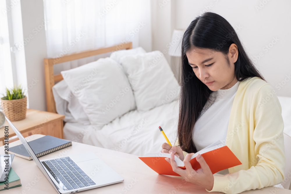 Poster asian female freelancer reading and jot down note on book while working with laptop on table in bedr