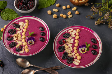 Healthy breakfast smoothie bowl with blackberries, gooseberries, peanuts, hazelnuts, decorated with flowers on a dark background, horizontal photo