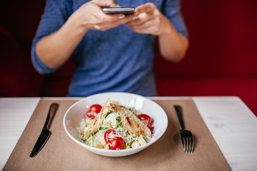 A woman in a blue blouse taking photo of fresh salad Caesar on the table with her smartphone. Social media concept