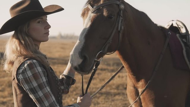 Blonde girl with long hair in cowboy hat stroking and hugging horse outdoors. Close up of beautiful young woman with her dark horse enjoying nature. Love and friendship concept.