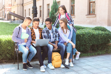 Young students with laptop sitting on bench outdoors