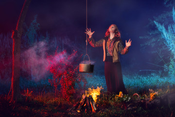 young witch by  fire in night forest prepares  magic potion