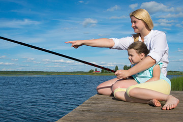 Mom holds the fishing rod and hand shows her daughter on the catch, on the pier, against the backdrop of a beautiful landscape, close-up, there is a place for an inscription