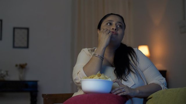 Overweight Indian woman taking handfull of chips and eating in front of a television - Junk food concept. A happy obese woman with a fat belly eating chips while watching television - Unhealthy lif...