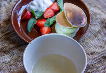 great breakfast. Fritters on plates with chopped strawberries and mint leaves. A plate and a cup of tea are on a jute napkin.