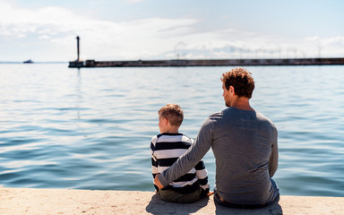 Rear view of father with small son sittting outdoors in town by the sea, talking.