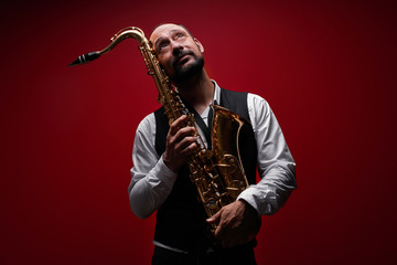 Fototapeta na wymiar Portrait of professional musician saxophonist man in suit plays jazz music on saxophone, red background in a photo studio