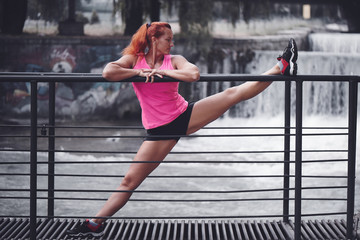 Adult red haired woman athlete in pink top and shorts performs morning stretching exercises at the river. Healthy lifestyle, yoga, fitness.