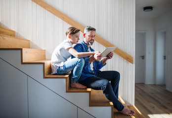 Mature father with small son sitting on the stairs indoors, using tablet.